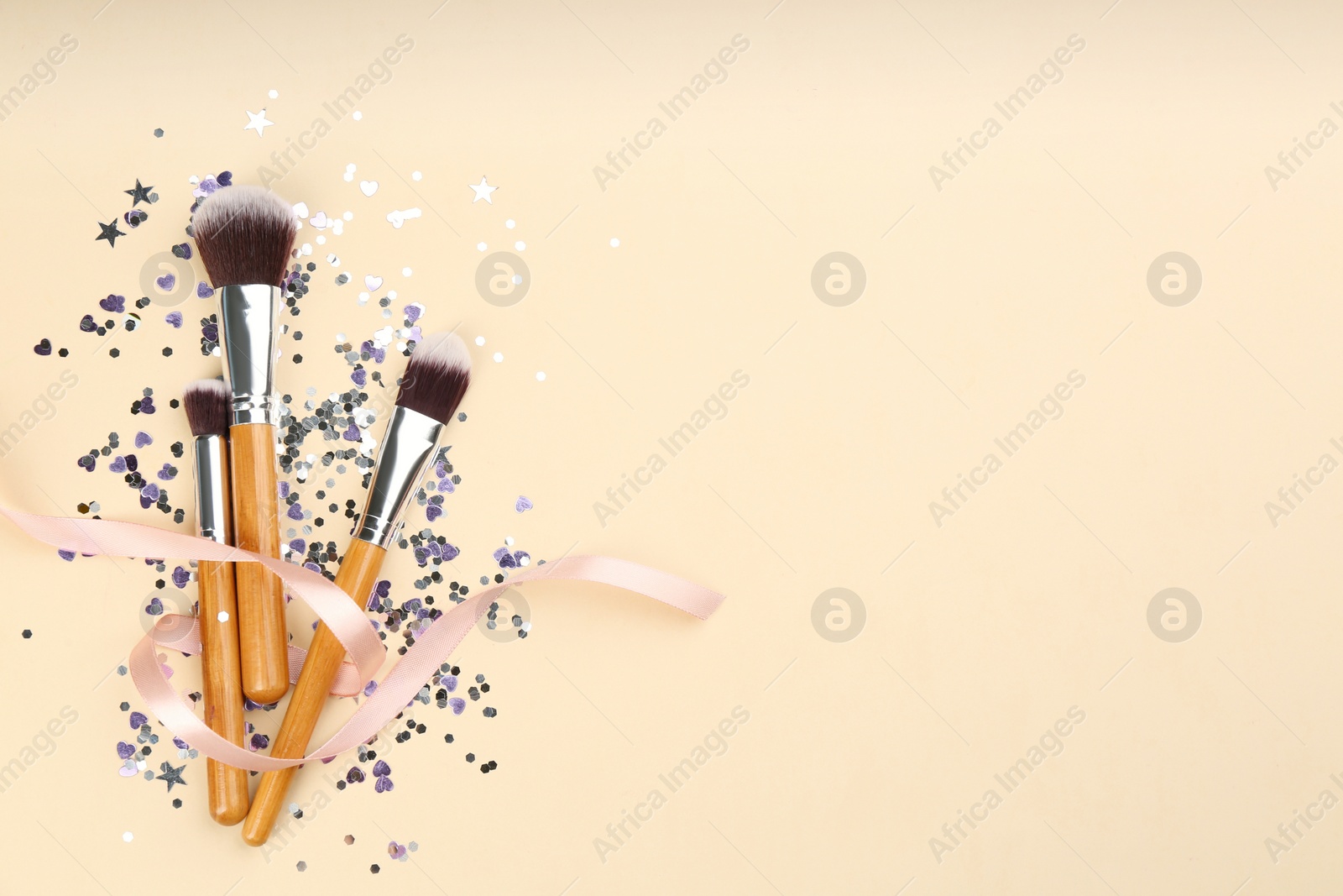 Photo of Different makeup brushes, ribbon and shiny confetti on beige background, flat lay. Space for text