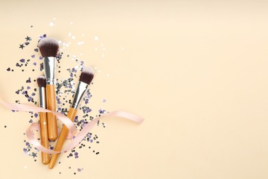 Photo of Different makeup brushes, ribbon and shiny confetti on beige background, flat lay. Space for text