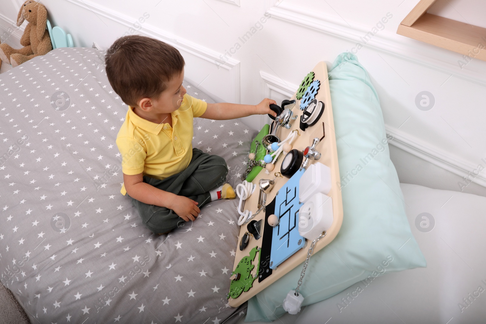 Photo of Little boy playing with busy board on bed at home, above view