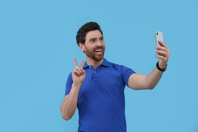Photo of Smiling man taking selfie with smartphone and showing peace sign on light blue background