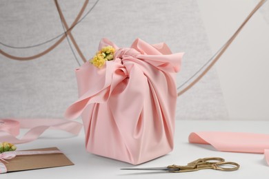 Photo of Furoshiki technique. Gift packed in pink fabric, card, flowers and scissors on white table