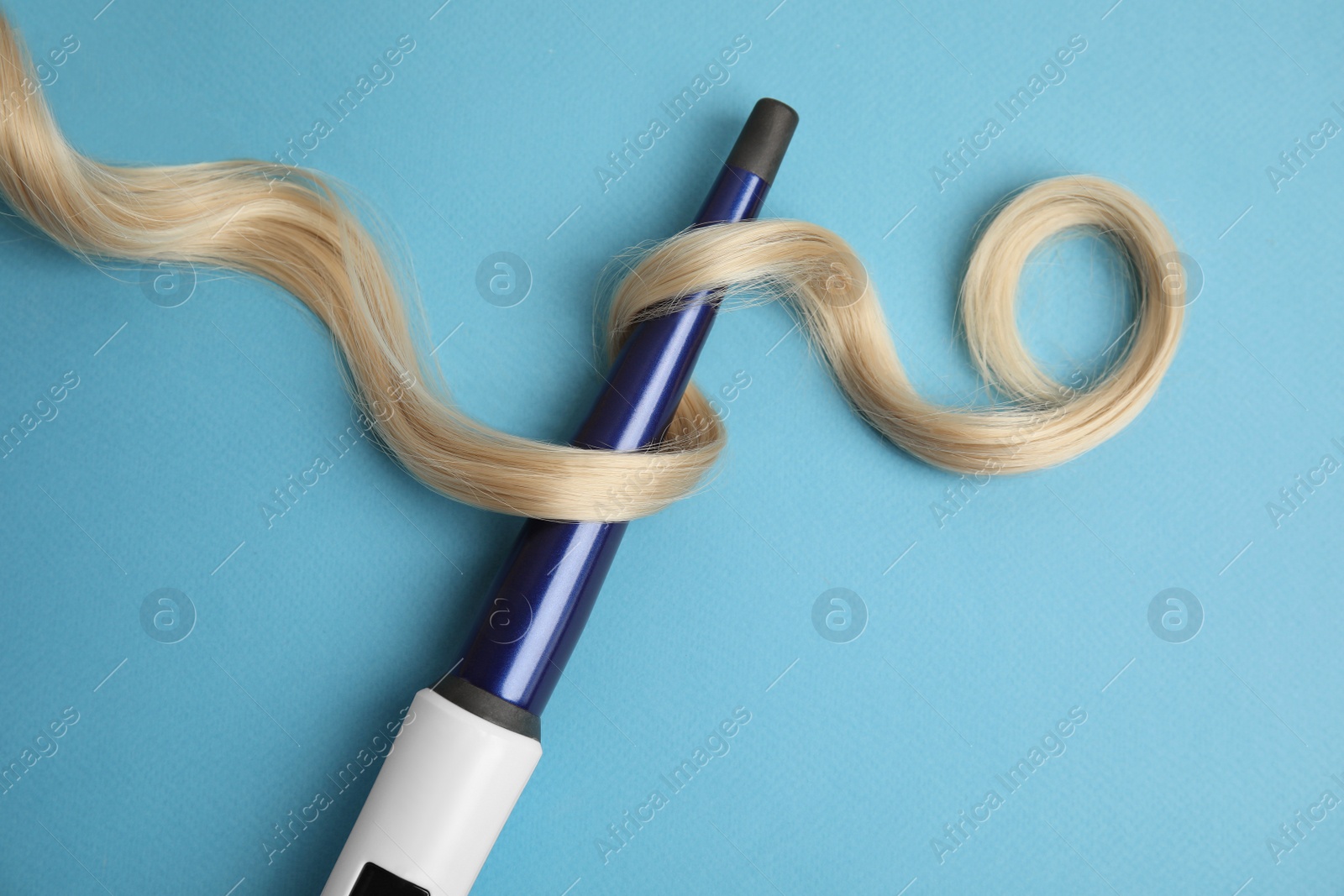 Photo of Curling iron with blonde hair lock on light blue background, flat lay