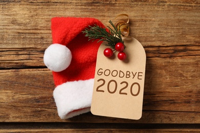 Tag with text Goodbye 2020 and Santa hat on wooden background, flat lay