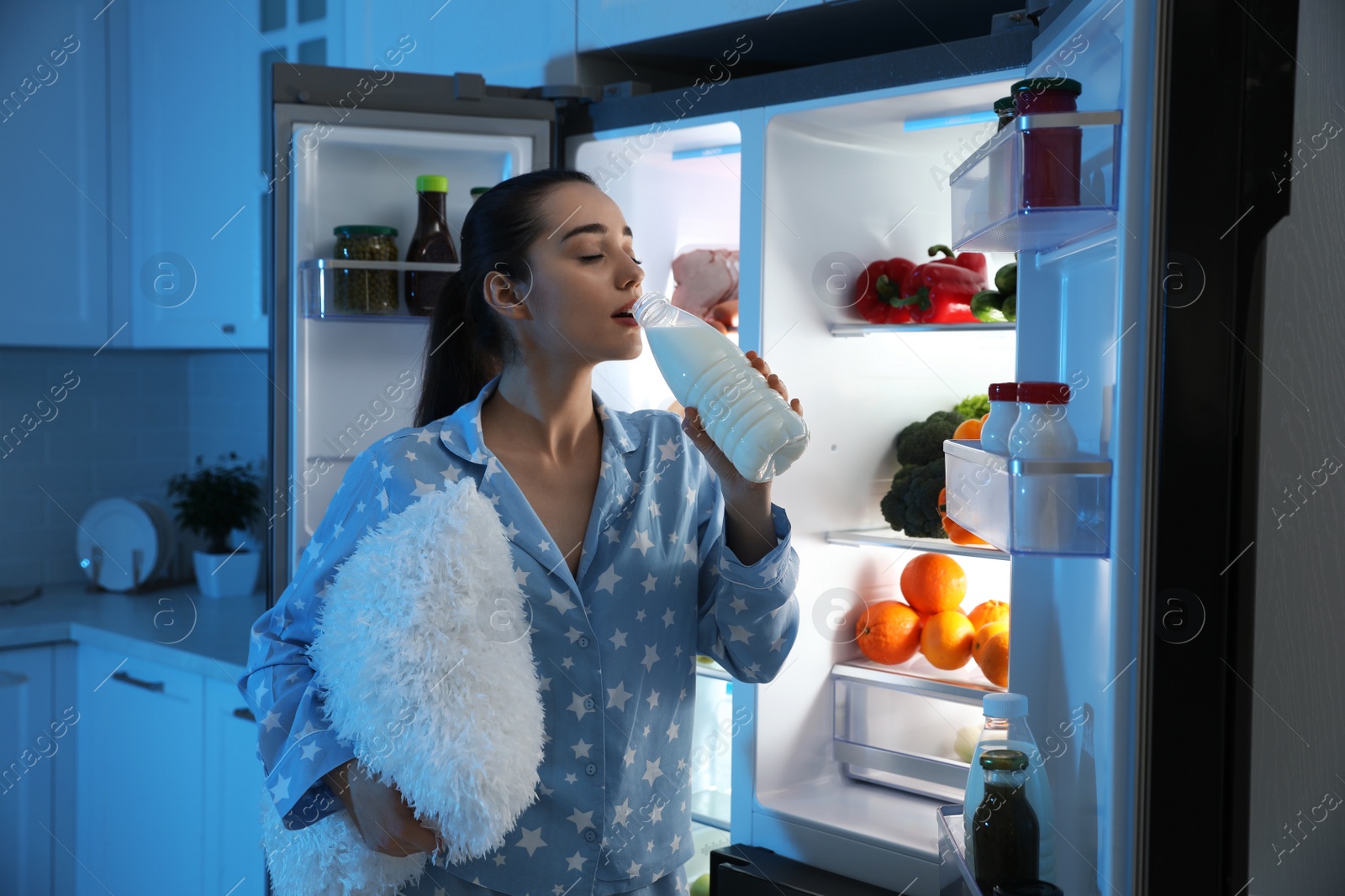 Photo of Young woman with pillow drinking milk near open refrigerator at night
