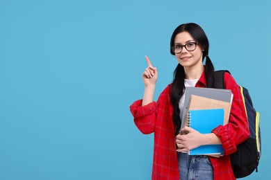 Smiling student with notebooks, folder and backpack pointing at something on light blue background. Space for text