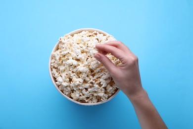 Woman taking delicious popcorn from paper bucket on light blue background, top view