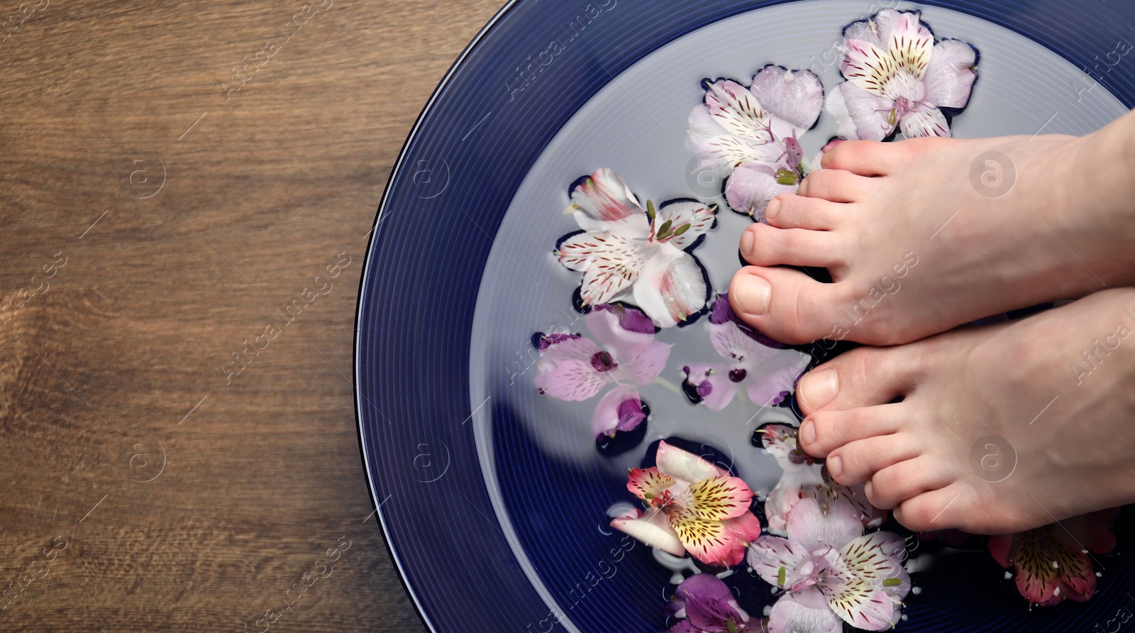 Photo of Woman putting her feet in bowl with water and flowers on wooden floor, top view with space for text. Spa treatment