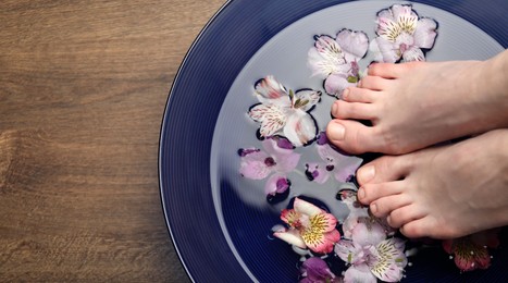 Woman putting her feet in bowl with water and flowers on wooden floor, top view with space for text. Spa treatment