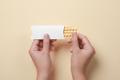 Woman holding blister with birth control pills on beige background, top view