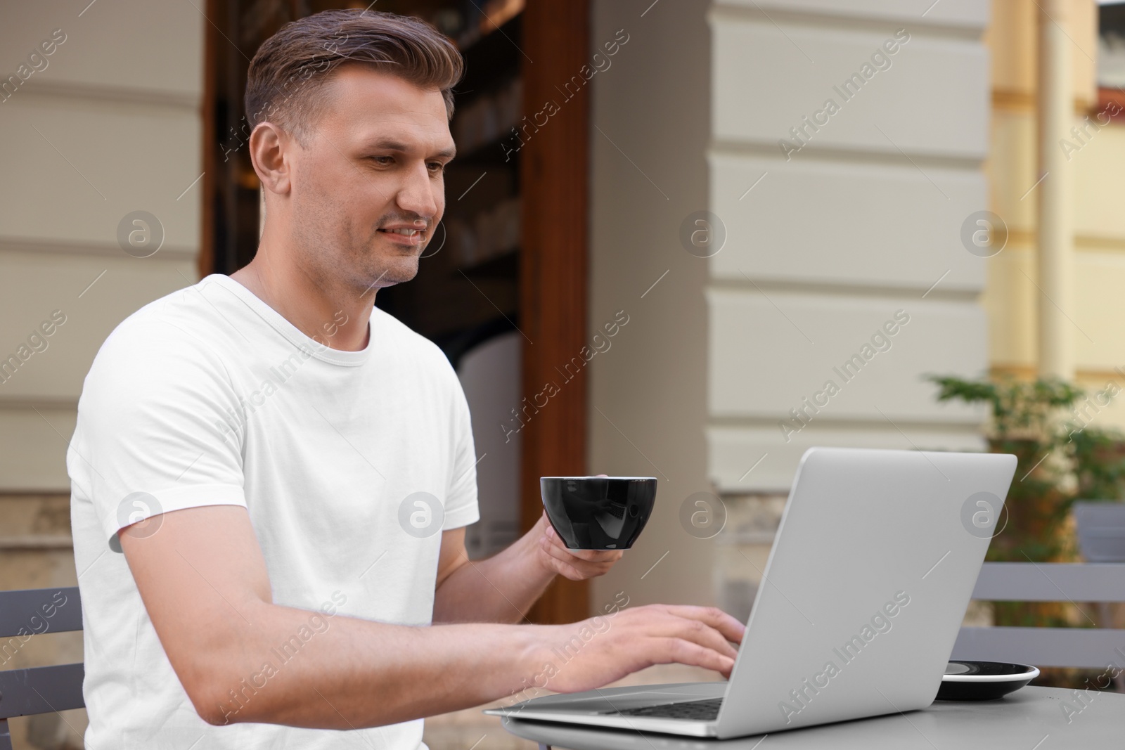 Photo of Handsome man with cup of drink working on laptop at table in outdoor cafe