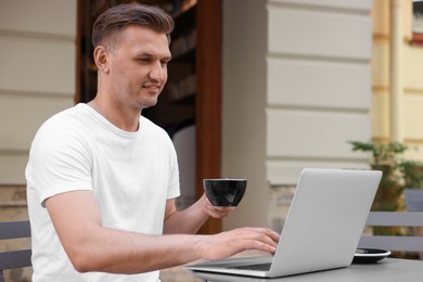 Handsome man with cup of drink working on laptop at table in outdoor cafe