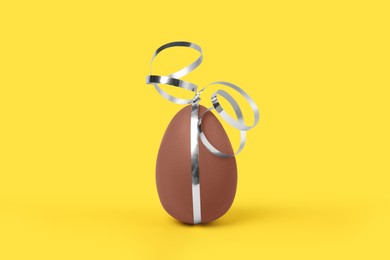 Delicious chocolate egg with silver ribbon on yellow background, closeup