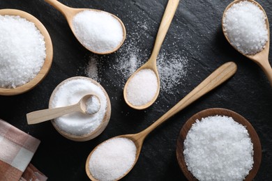 Photo of Organic white salt in bowls and spoons on black table, flat lay