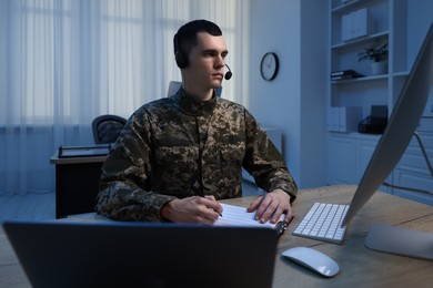 Photo of Military service. Young soldier in headphones working at wooden table in office at night