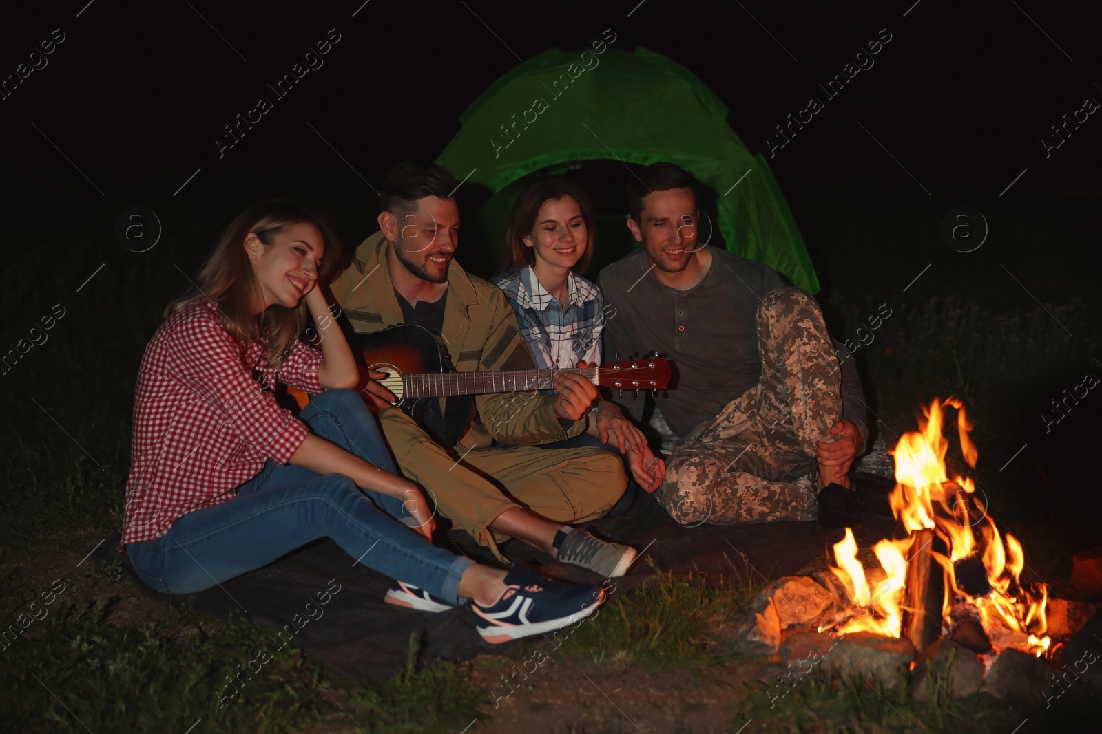 Photo of Young man playing guitar for friends near bonfire at night. Camping season