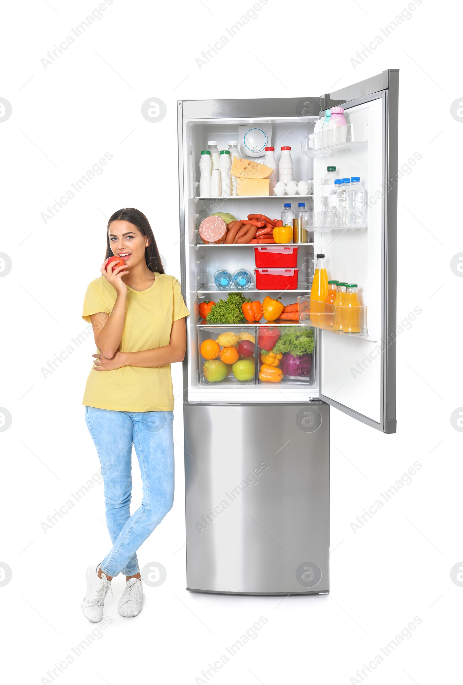 Photo of Young woman eating apple near open refrigerator on white background