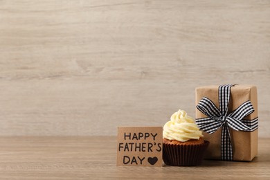 Photo of Card with phrase Happy Father's Day, gift box and tasty cupcake on wooden background, space for text