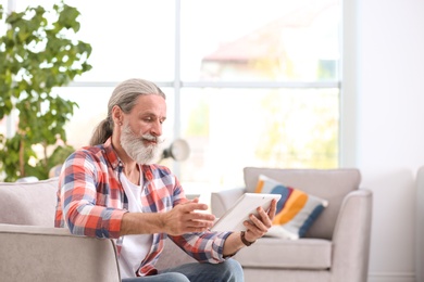 Handsome mature man with tablet sitting in armchair indoors