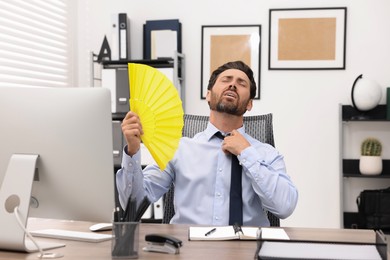 Bearded businessman waving yellow hand fan to cool himself at table in office