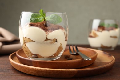 Delicious tiramisu in glass, mint leaves, coffee beans and fork on wooden table, closeup