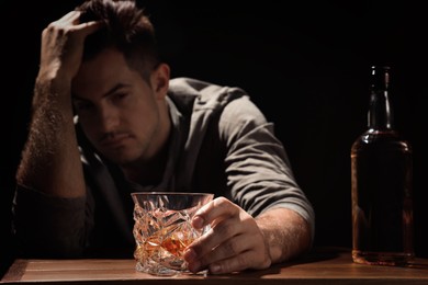 Photo of Addicted man at wooden table against black background, focus on glass of alcoholic drink