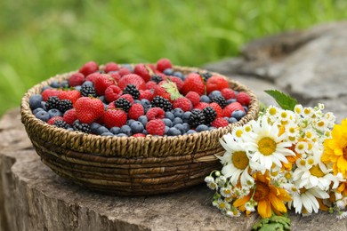 Wicker bowl with different fresh ripe berries and beautiful flowers on wooden surface outdoors