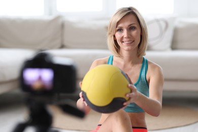 Smiling sports blogger holding medicine ball while recording fitness lesson with camera at home