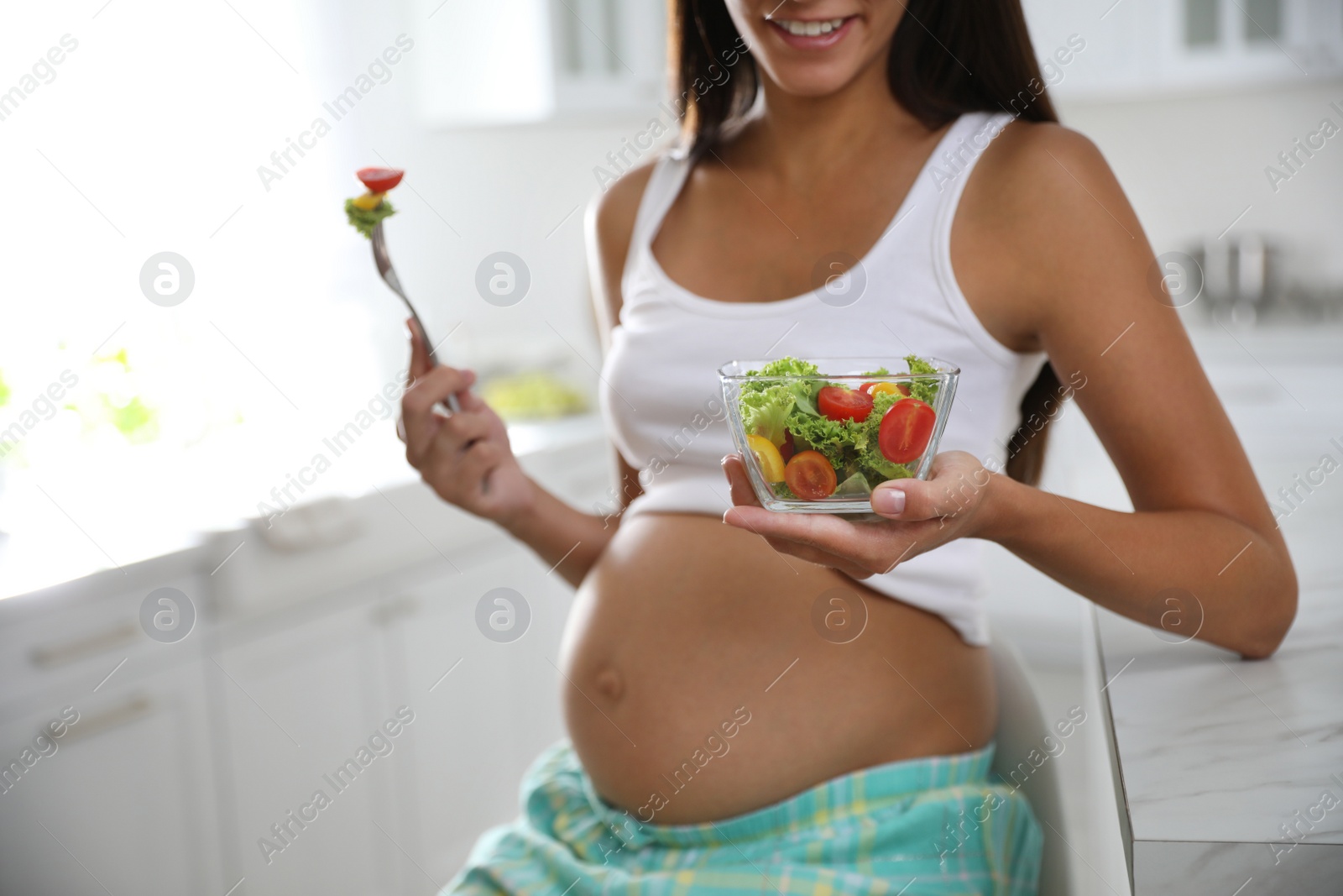 Photo of Young pregnant woman with bowl of vegetable salad at table in kitchen, closeup. Taking care of baby health