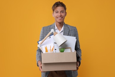 Happy unemployed young man with box of personal office belongings on orange background
