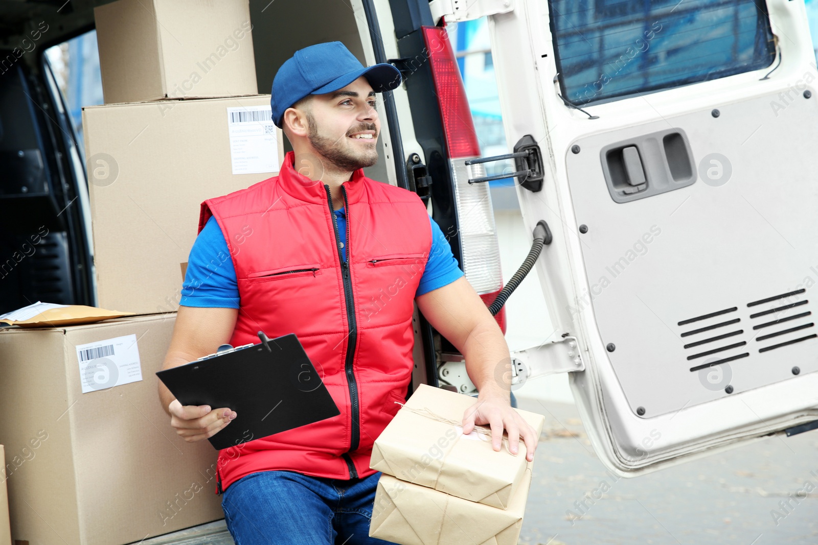 Photo of Young courier with parcels and clipboard near delivery van outdoors