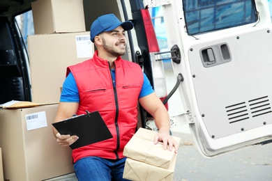 Photo of Young courier with parcels and clipboard near delivery van outdoors