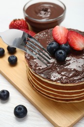 Eating tasty pancakes with chocolate paste and berries with fork on white wooden table, closeup