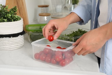 Photo of Man putting cherry tomato into plastic container with fresh vegetables in kitchen, closeup. Food storage
