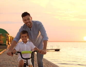Happy father teaching his son to ride bicycle near river at sunset