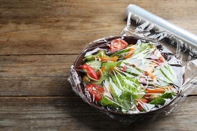 Photo of Bowl of fresh salad with plastic food wrap on wooden table. Space for text