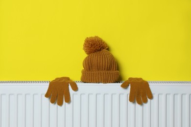Photo of Knitted hat and gloves on heating radiator near yellow wall