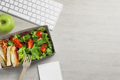 Photo of Container of tasty food, keyboard, apple and fork on white wooden table, flat lay with space for text. Business lunch