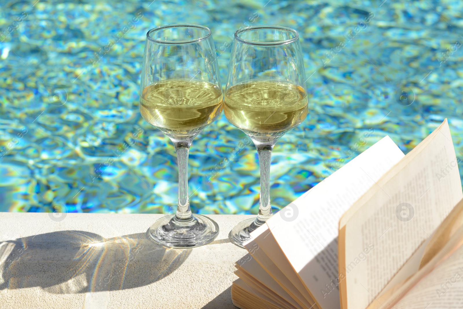 Photo of Glasses of tasty wine and open book on swimming pool edge