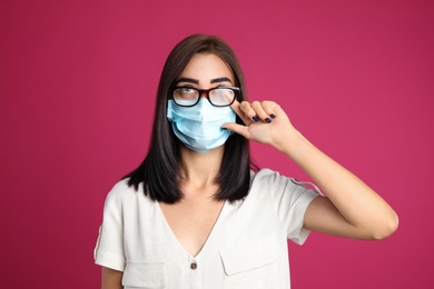 Photo of Young woman wiping foggy glasses caused by wearing disposable mask on pink background. Protective measure during coronavirus pandemic