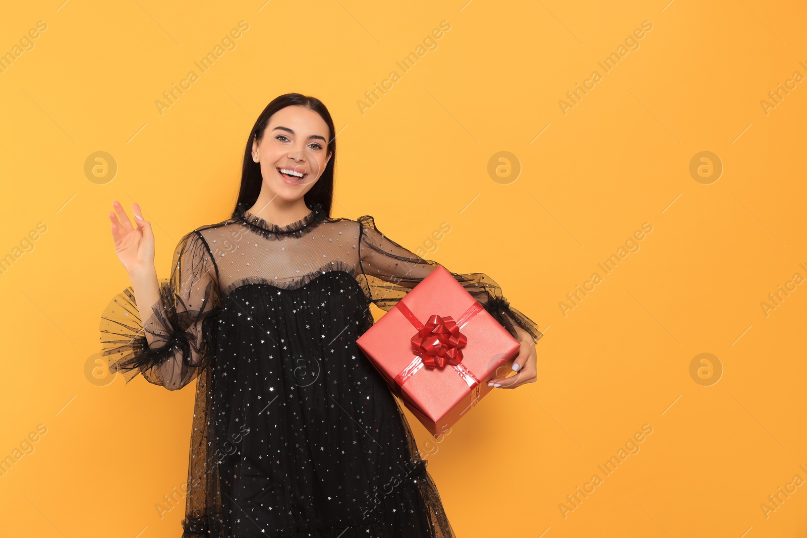 Photo of Happy young woman in festive dress with gift box on orange background, space for text