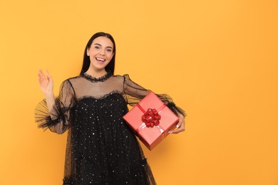 Happy young woman in festive dress with gift box on orange background, space for text