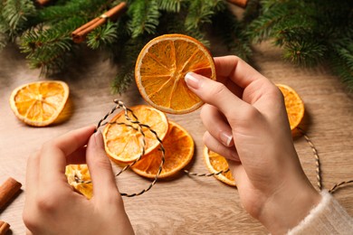 Photo of Woman making handmade garland from dry orange slices at wooden table, closeup