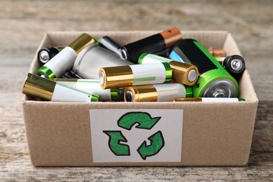 Used batteries in cardboard box with recycling symbol on wooden table, closeup
