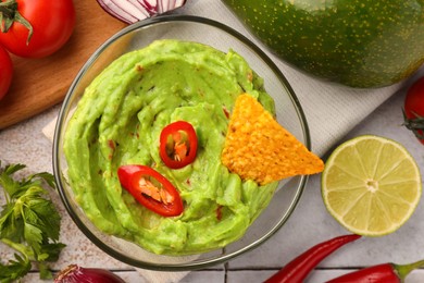 Bowl of delicious guacamole, nachos chip and ingredients on white tiled table, flat lay