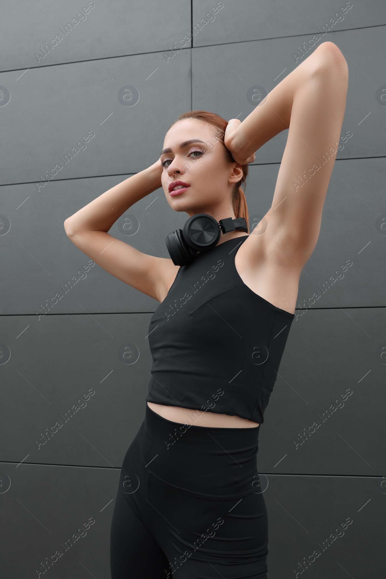 Photo of Beautiful woman in stylish gym clothes with headphones posing near dark grey wall on street