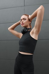 Beautiful woman in stylish gym clothes with headphones posing near dark grey wall on street