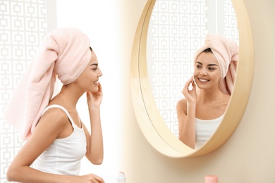 Photo of Pretty young woman with towel on head near mirror in bathroom