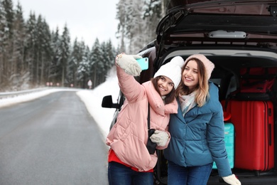 Photo of Friends taking selfie near open car trunk full of luggage on road, space for text. Winter vacation