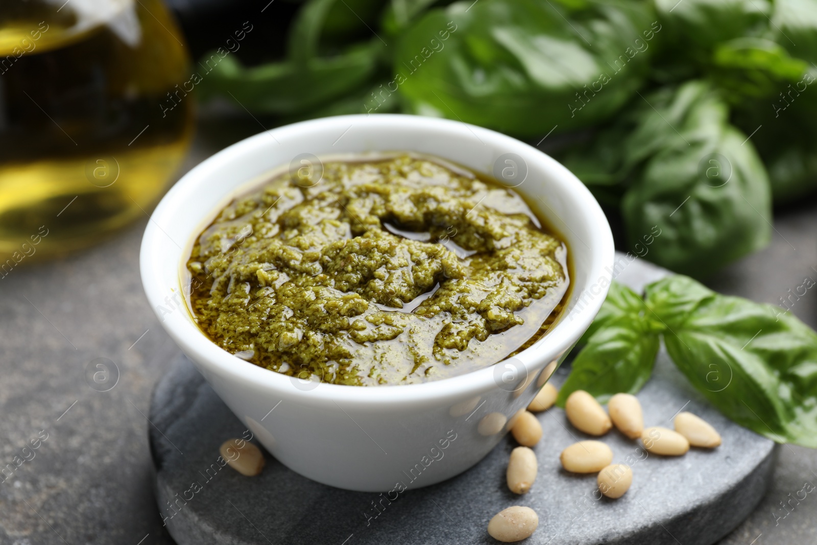 Photo of Tasty pesto sauce in bowl, basil and pine nuts on grey table, closeup