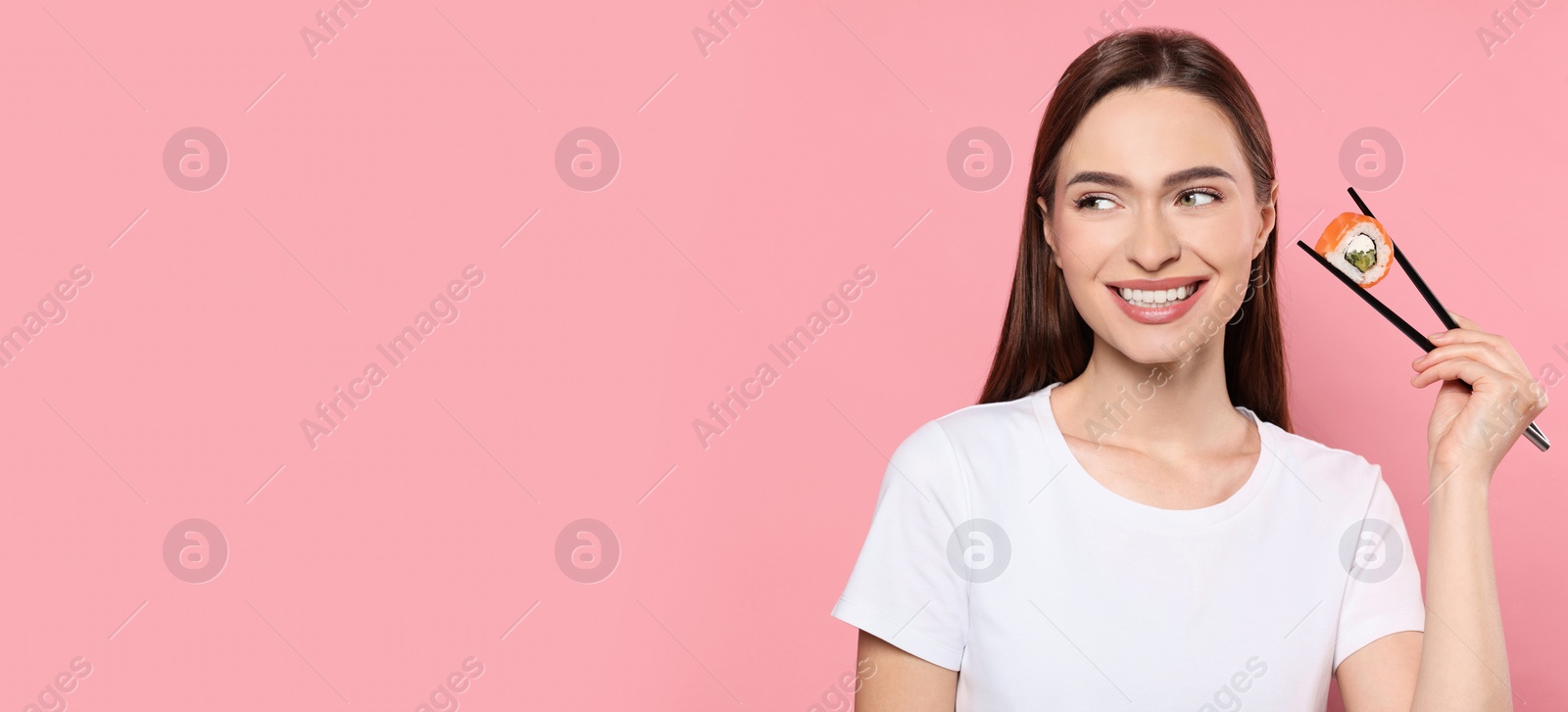 Photo of Beautiful young woman holding sushi roll with chopsticks on pink background. Space for text